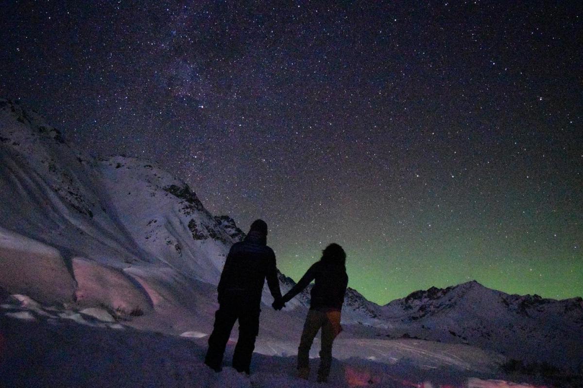 Two people holding hands under a starry sky, with a faint green aurora over snowy mountains